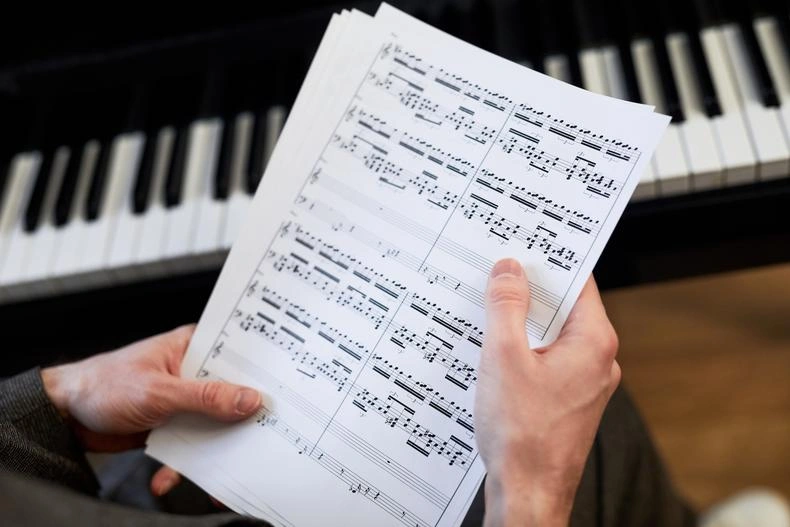 Hands holding sheet music with a grand piano keyboard visible in the background.